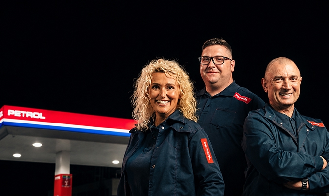 Na sliki so trije zaposleni v delovnih oblačilih Petrol, fotografirani pred Petrolovim bencinskim servisom. V ozadju je viden osvetljen nadstrešek bencinskega servisa, prizor pa ponazarja delo, sodelovanje in karierne priložnosti v podjetju Petrol.