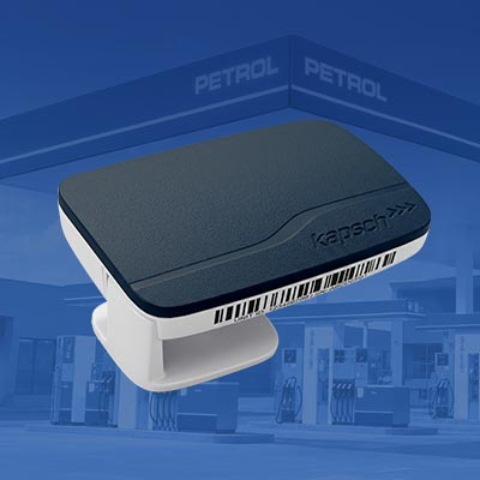 Na slici je uređaj za elektronsku identifikaciju vozila, prikazan na plavoj pozadini sa Petrol benzinskom stanicom u pozadini. Uređaj je crne boje, pravougaonog oblika sa zaobljenim ivicama i postavljen na beli nosač. Prikaz ilustruje opremu za automatsku identifikaciju vozila i beskontaktno korišćenje usluga na Petrol prodajnim mestima.