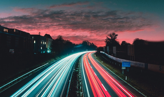 Na slici je višetračna saobraćajnica u večernjim satima, kojom se odvija saobraćaj u oba smera. Zbog duge ekspozicije vidljivi su svetlosni tragovi vozila, dok se u pozadini nalaze zalazak sunca i objekti pored puta, što simbolizuje mobilnost, povezanost i saobraćajnu infrastrukturu.