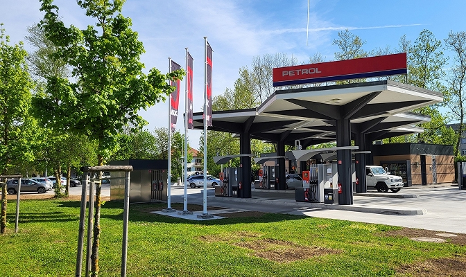 Na slici je Petrol benzinska stanica sa više točionih mesta za gorivo, parking prostorima i pratećim objektima. Oko stanice su vidljive Petrol zastave, zelene površine i drveće, a prizor simbolizuje širenje maloprodajne mreže i dostupnost usluga za vozače i putnike.